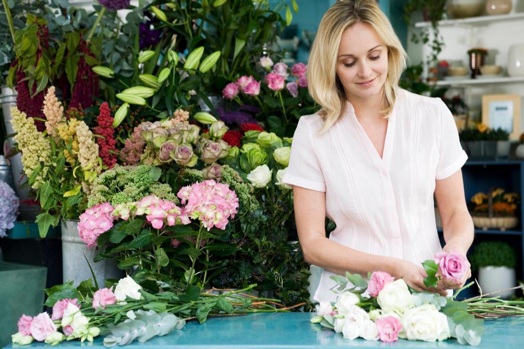 Eine junge Floristin steht an einem Tisch und bindet einen Strauß aus weißen und rosanen Rosen. Hinter ihr sind Behälter mit verschiedenen Blumen aufgebaut.