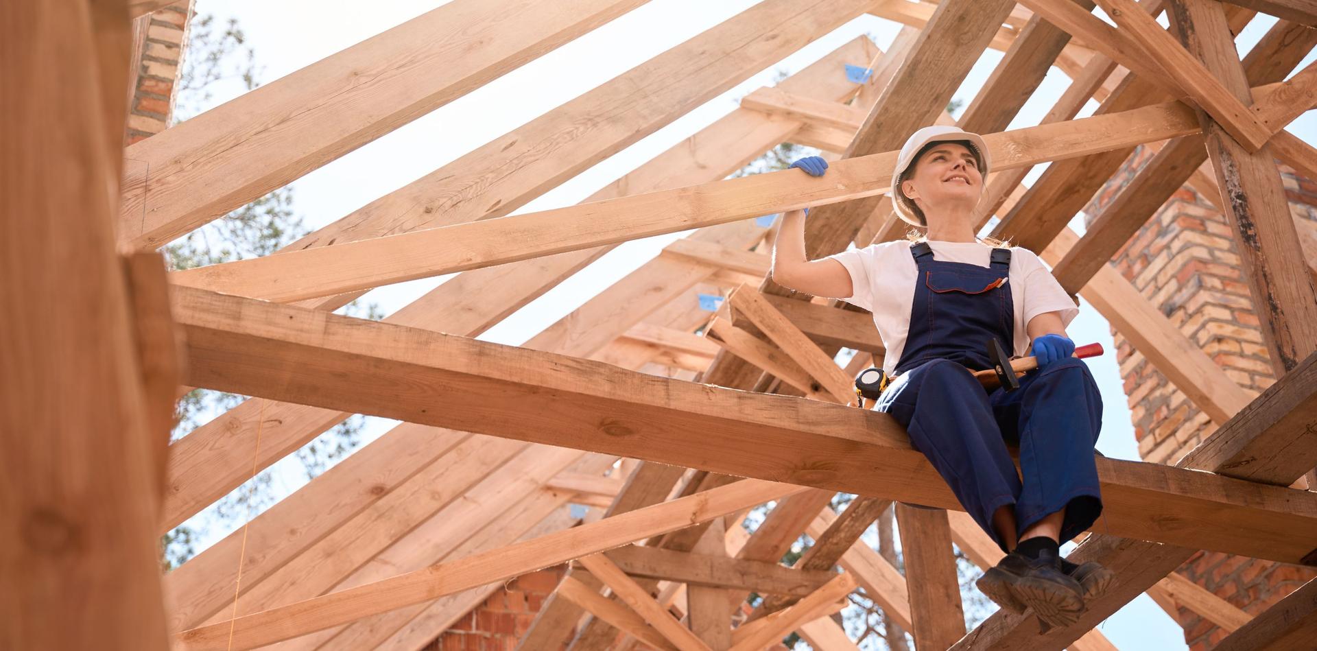 Eine Zimmerin sitzt mit Schutzhelm auf den Holzbalken eines im Bau befindlichen Dachstuhls und hält dabei einen Hammer in der Hand.