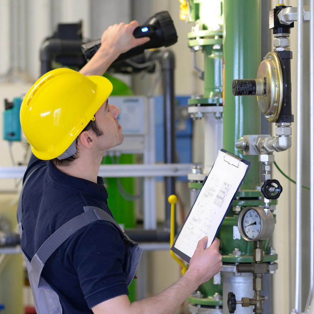 Ein Anlagenmechaniker SHK mit gelbem Schutzhelm und blauem Arbeitshemd scannt mit einer Lampe eine grüne Sanitär-Heizungs-Anlage ab und hält eine Klemmtafel mit Papieren in der Hand. Im industriellen Hintergrund Rohre, Ventile, Manometer und Kabel.