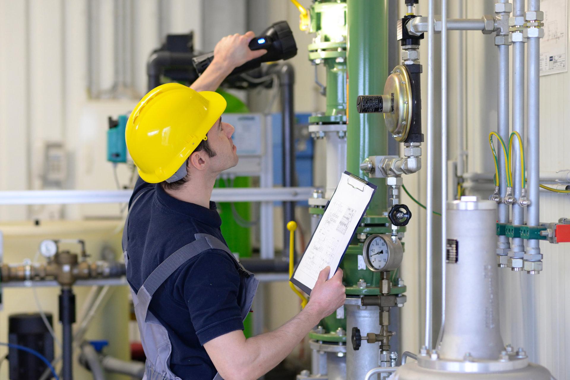 Ein Anlagenmechaniker SHK mit gelbem Schutzhelm und blauem Arbeitshemd scannt mit einer Lampe eine grüne Sanitär-Heizungs-Anlage ab und hält eine Klemmtafel mit Papieren in der Hand. Im industriellen Hintergrund Rohre, Ventile, Manometer und Kabel.