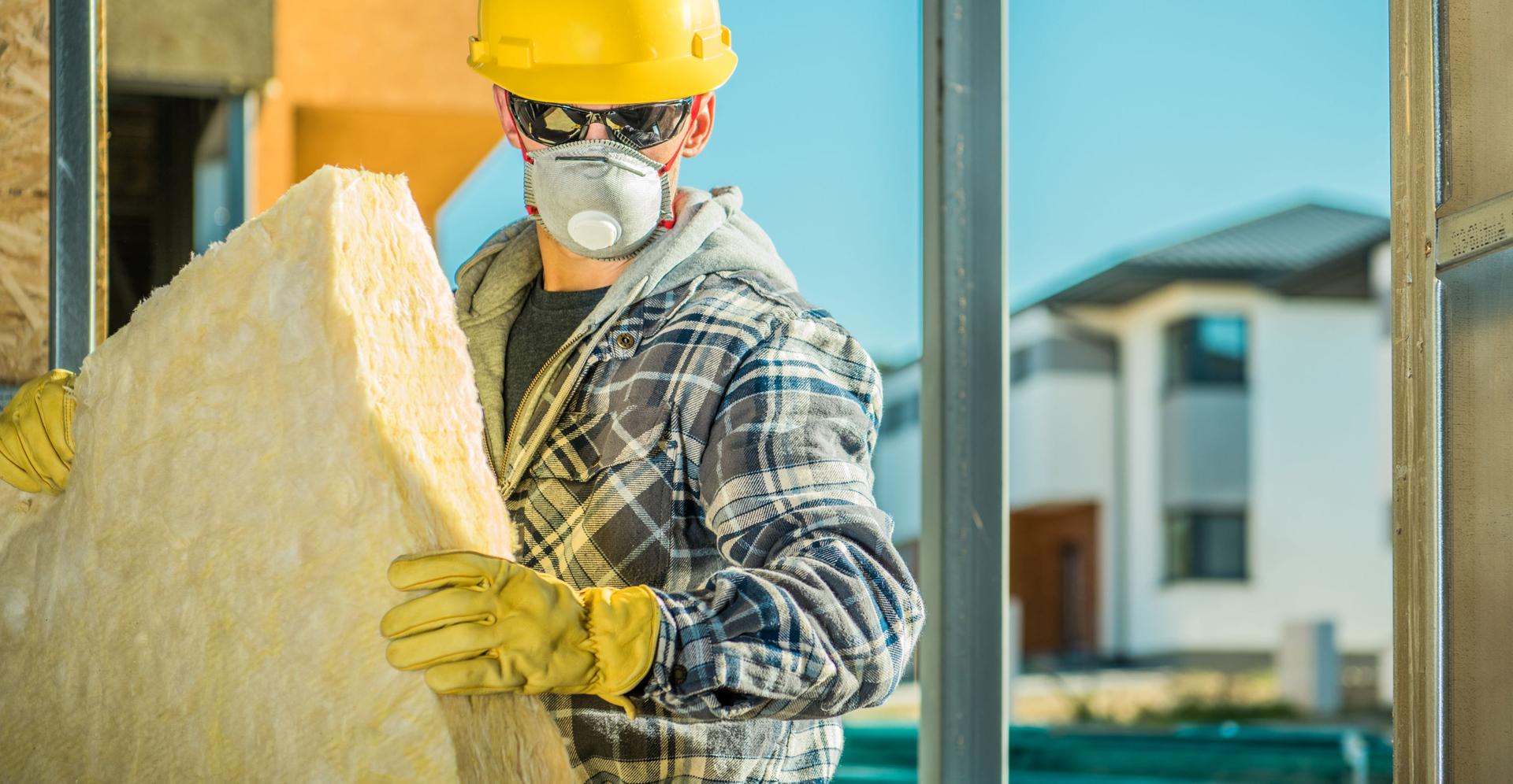 Eine Fachkraft mit Schutzkleidung, Helm und Atemmaske trägt auf einer Baustelle eine große Dämmplatte in den Händen.