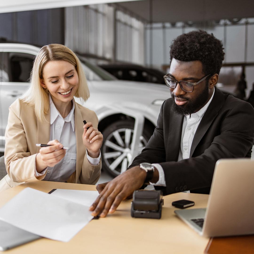 Eine blonde Automobilkauffrau in hellem Blazer und ein schwarzer Automobilkaufmann in Anzug beraten sich lachend am Schreibtisch mit Papieren, Kartenleser und Laptop in einer Autohaus-Verkaufsfläche neben einem silbernen SUV.