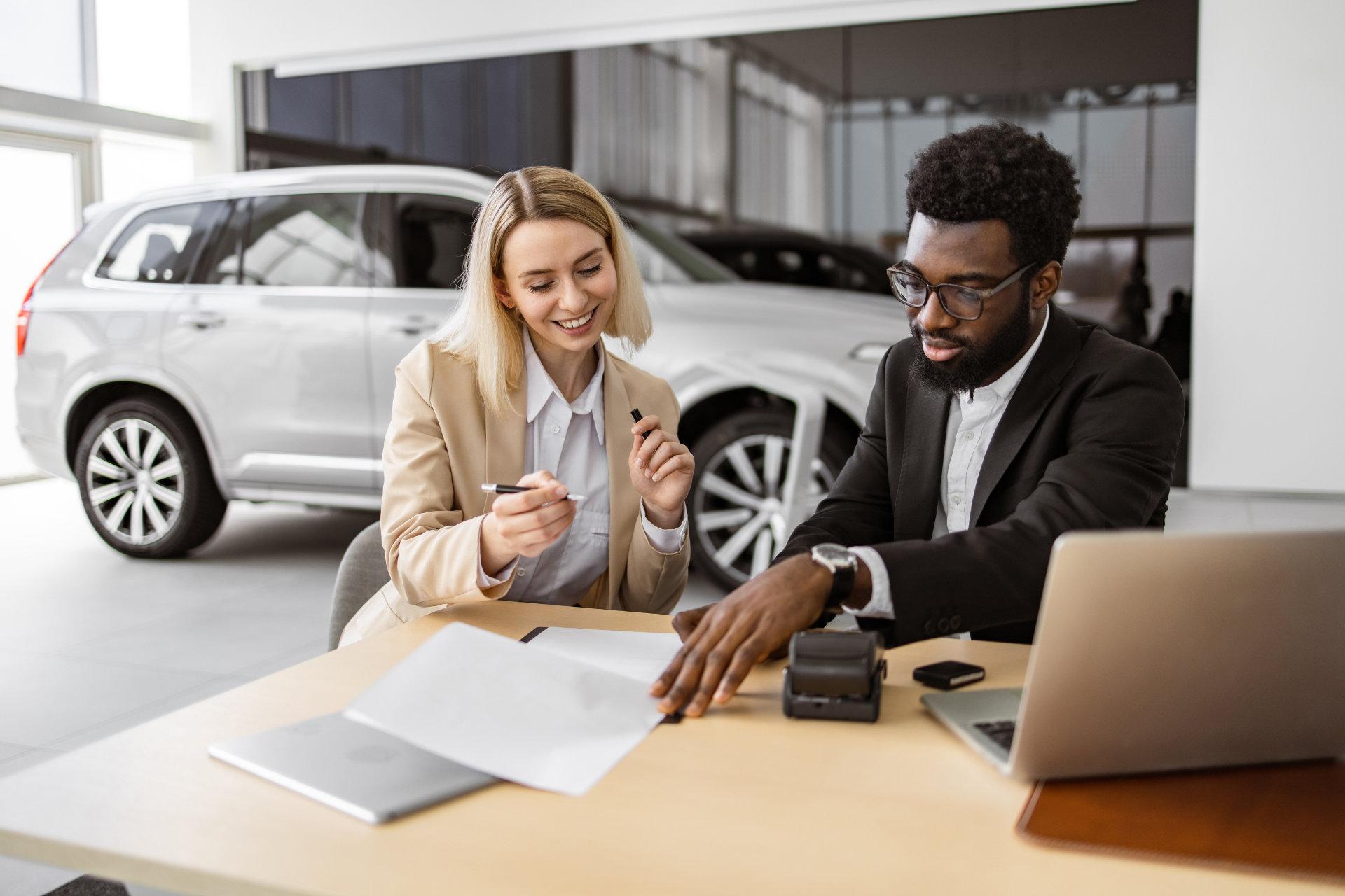 Eine blonde Automobilkauffrau in hellem Blazer und ein schwarzer Automobilkaufmann in Anzug beraten sich lachend am Schreibtisch mit Papieren, Kartenleser und Laptop in einer Autohaus-Verkaufsfläche neben einem silbernen SUV.