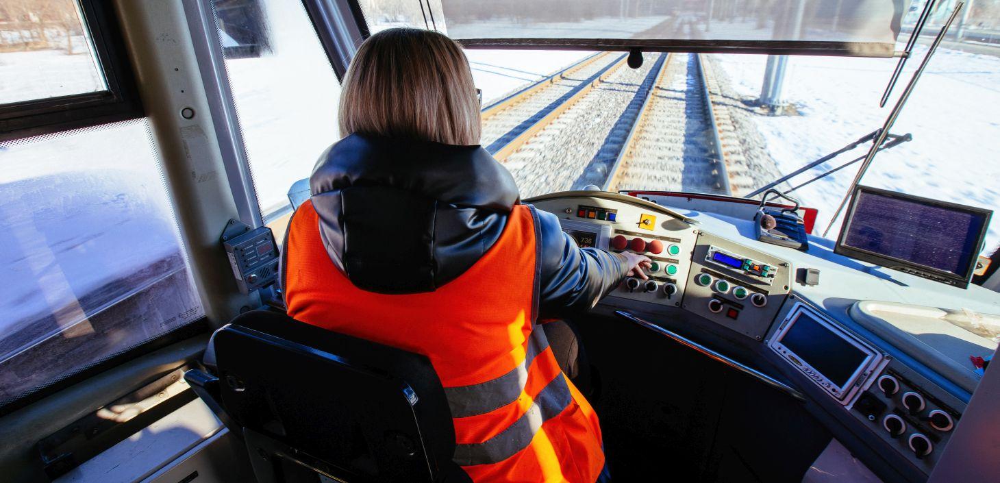 Eine junge Frau sitzt im Führerhaus einer Bahn. Durch die Scheibe vor ihr erkennt man die Gleise. Sie drückt auf einen Knopf.