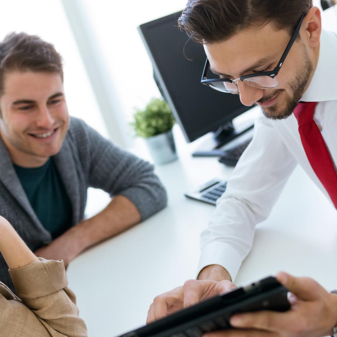 Ein Banker mit Brille, weißem Hemd und roter Krawatte zeigt einem lachenden Paar (junge Frau mit Brille in beigem Blazer und junger Mann in grauem Pullover) etwas auf einem Tablet während einer Beratung am weißen Schreibtisch in einer Bankfiliale. Im Hintergrund ein Computermonitor und eine Topfpflanze.