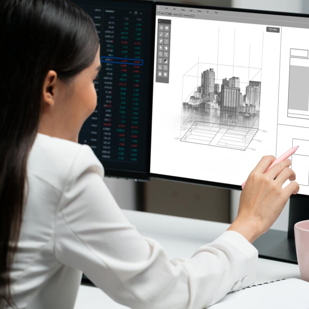Eine junge asiatische Bauzeichnerin mit langem schwarzem Haar in weißer Bluse arbeitet von hinten an einem großen Monitor mit 3D-Stadtmodell und Zeichnungstools, zeichnet mit Stift nach und hat eine rosa Tasse sowie Notizblock auf dem Tisch sowie einen zweiten Monitor daneben.