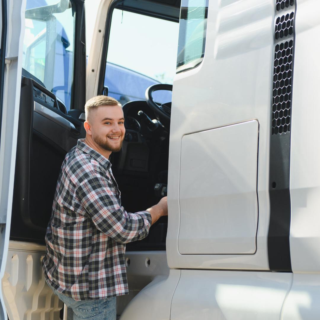 Ein junger Berufskraftfahrer mit kurzem hellen Haar und Karohemd lehnt lachend aus der geöffneten Fahrertür eines weißen Lkw mit Anhänger und greift hinein.