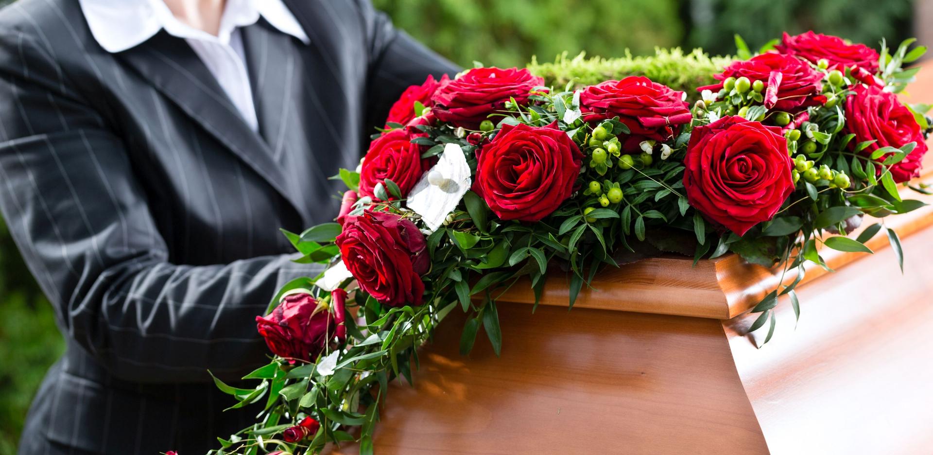 Eine Frau positioniert ein Blumengesteck auf einem Sarg. Das gesteck besteht überwiegend aus roten Rosen.