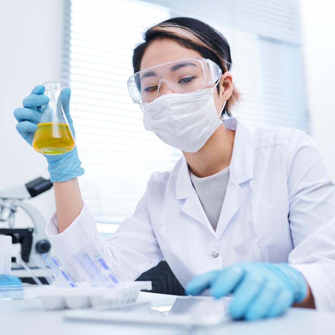 Eine junge asiatische Chemielaborantin mit Brille, weißer Maske und Pferdeschwanz im weißen Laborkittel hält mit blauen Handschuhen einen Erlenmeyerkolben mit gelber Flüssigkeit hoch und betrachtet sie. Auf dem Labortisch stehen Reagenzglasständer mit blauen Flüssigkeiten, ein Mikroskop, Pipetten und weitere Gefäße in einem hellen Laborraum mit Fenster.
