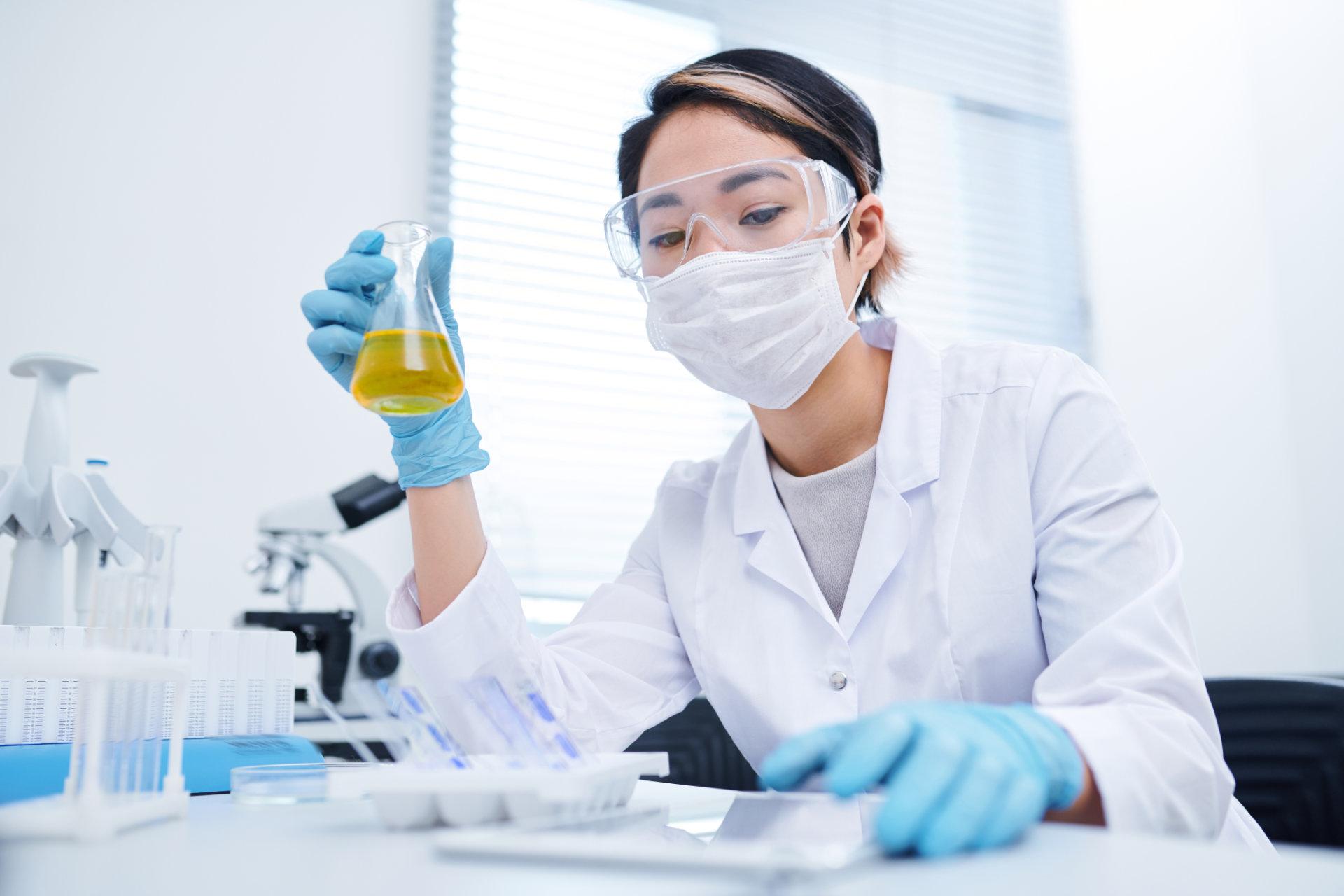 Eine junge asiatische Chemielaborantin mit Brille, weißer Maske und Pferdeschwanz im weißen Laborkittel hält mit blauen Handschuhen einen Erlenmeyerkolben mit gelber Flüssigkeit hoch und betrachtet sie. Auf dem Labortisch stehen Reagenzglasständer mit blauen Flüssigkeiten, ein Mikroskop, Pipetten und weitere Gefäße in einem hellen Laborraum mit Fenster.
