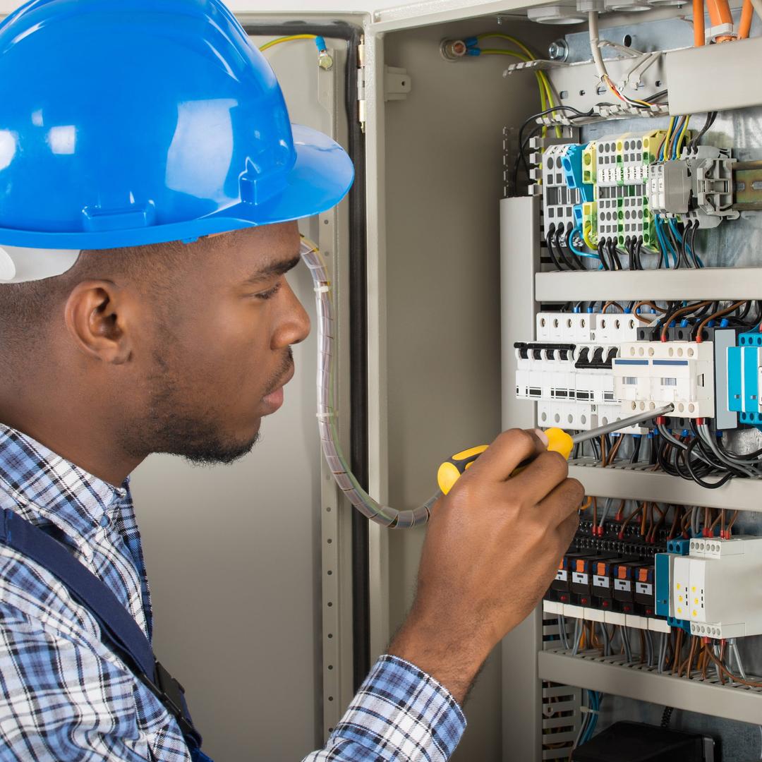 Ein Elektriker mit blauem Schutzhelm arbeitet mit einem Schraubendreher konzentriert an den ordentlich angeordneten Schaltern und Kabeln in einem geöffneten Schaltschrank.