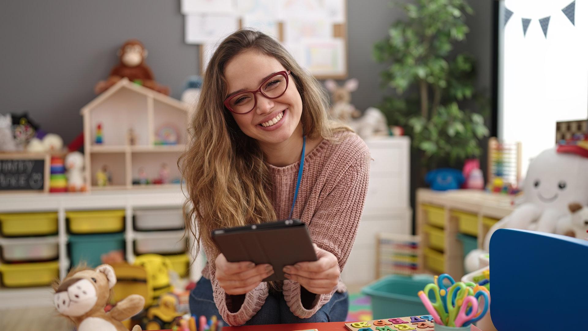 Eine lachende Erzieherin mit Brille, welligen Haaren und beigem Pullover hält ein Tablet in einem bunt eingerichteten Kindergartenraum mit Spielzeug, Puppenhaus, Teddybären, Regalen und Girlande.