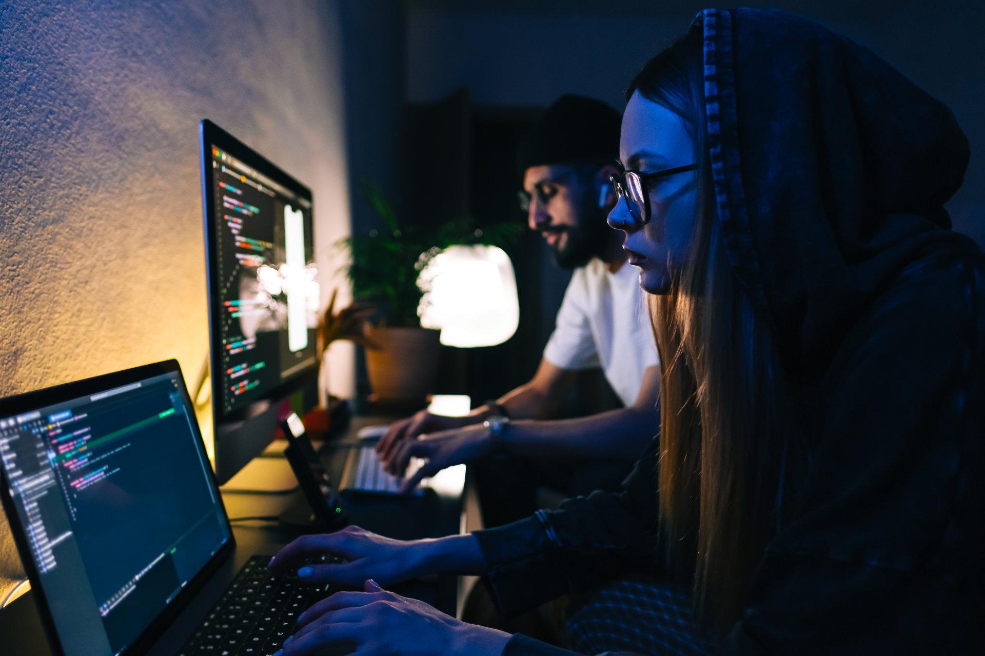Zwei Fachinformatiker für Anwendungsentwicklung sitzen in einem dunklen Büro und schauen sich etwas auf ihrem Computer an.