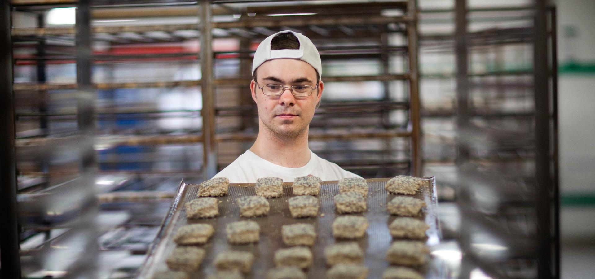 Ein junger Mann, vermutlich ein Fachpraktiker im Bäckerhandwerk, holt ein Blech voller Backwaren aus einem Rollwagen oder schiebt es gerade hinein. Es scheint sich bei den Backwaren um Körnerbrötchen zu handeln.