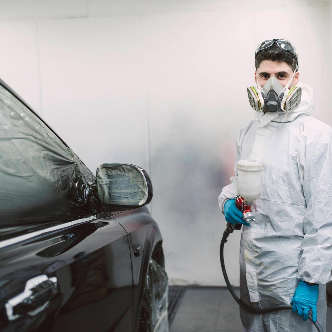 Ein junger Fahrzeuglackierer mit Schutzbrille und Atemschutzmaske in weißem Maleranzug und blauen Handschuhen lackiert mit Spritzpistole ein schwarzes Auto in einer Lackierkabine.