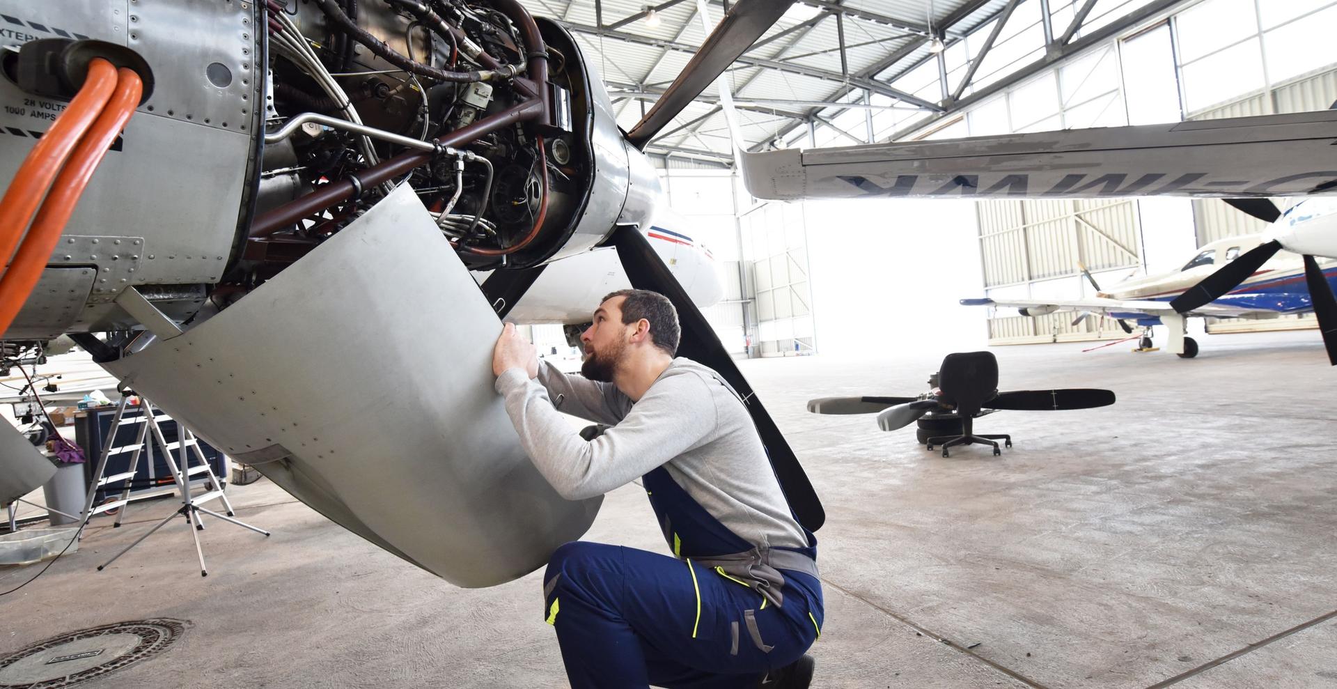 Ein Mechaniker kniet in einer Flugzeughalle und überprüft konzentriert den geöffneten Motor eines kleinen Flugzeugs.