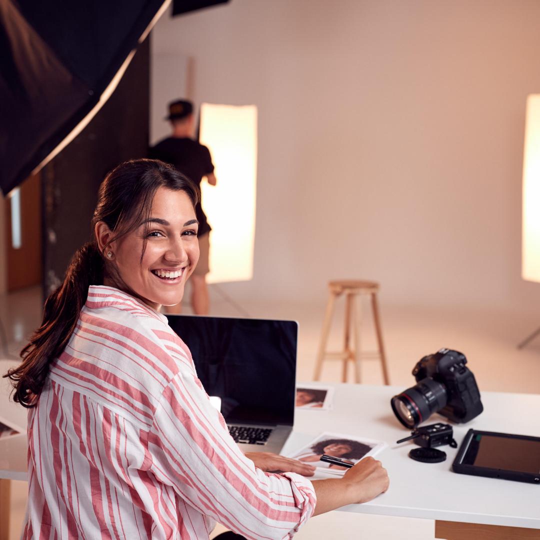 Eine Frau befindet sich in einem Fotostudio und lächelt mit einer Kamera und einem Laptop vor ihr auf dem Tisch in die Kamera.