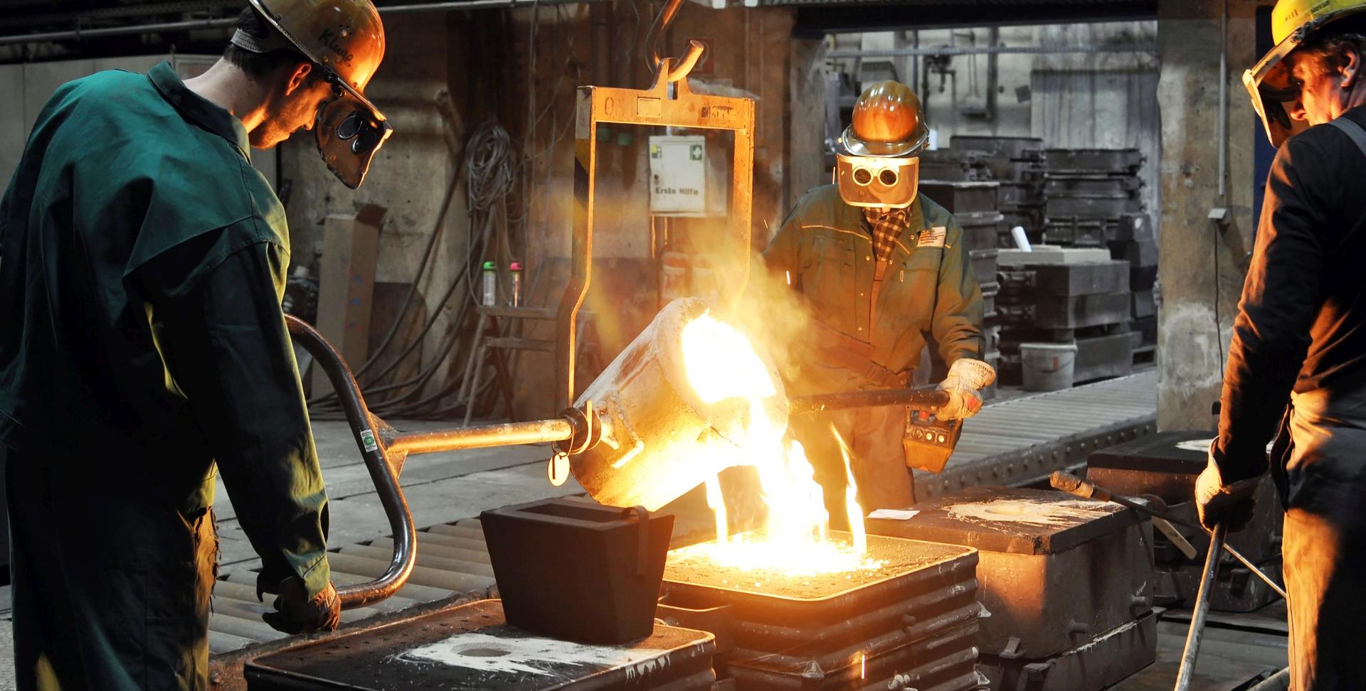 Das Bild zeigt mehrere Gießereimechaniker in Schutzkleidung und mit Schutzhelmen, die in einer Gießerei flüssiges, glühendes Metall aus einem Schmelzgefäß in eine Form gießen, um Metallbauteile herzustellen.