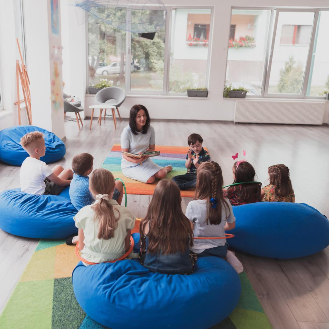 Eine Heilerziehungspflegerin sitzt mit mehreren Kindern auf Sitzsäcken in einem großen hellen Raum.