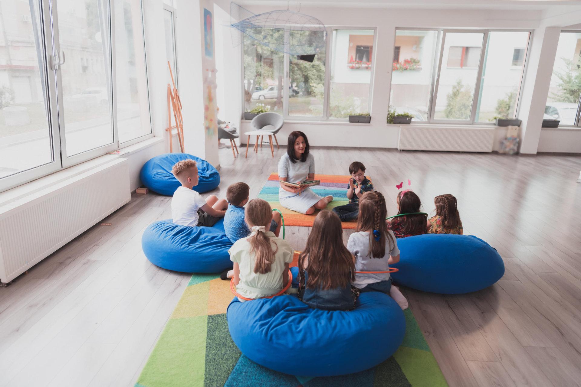Eine Heilerziehungspflegerin sitzt mit mehreren Kindern auf Sitzsäcken in einem großen hellen Raum.