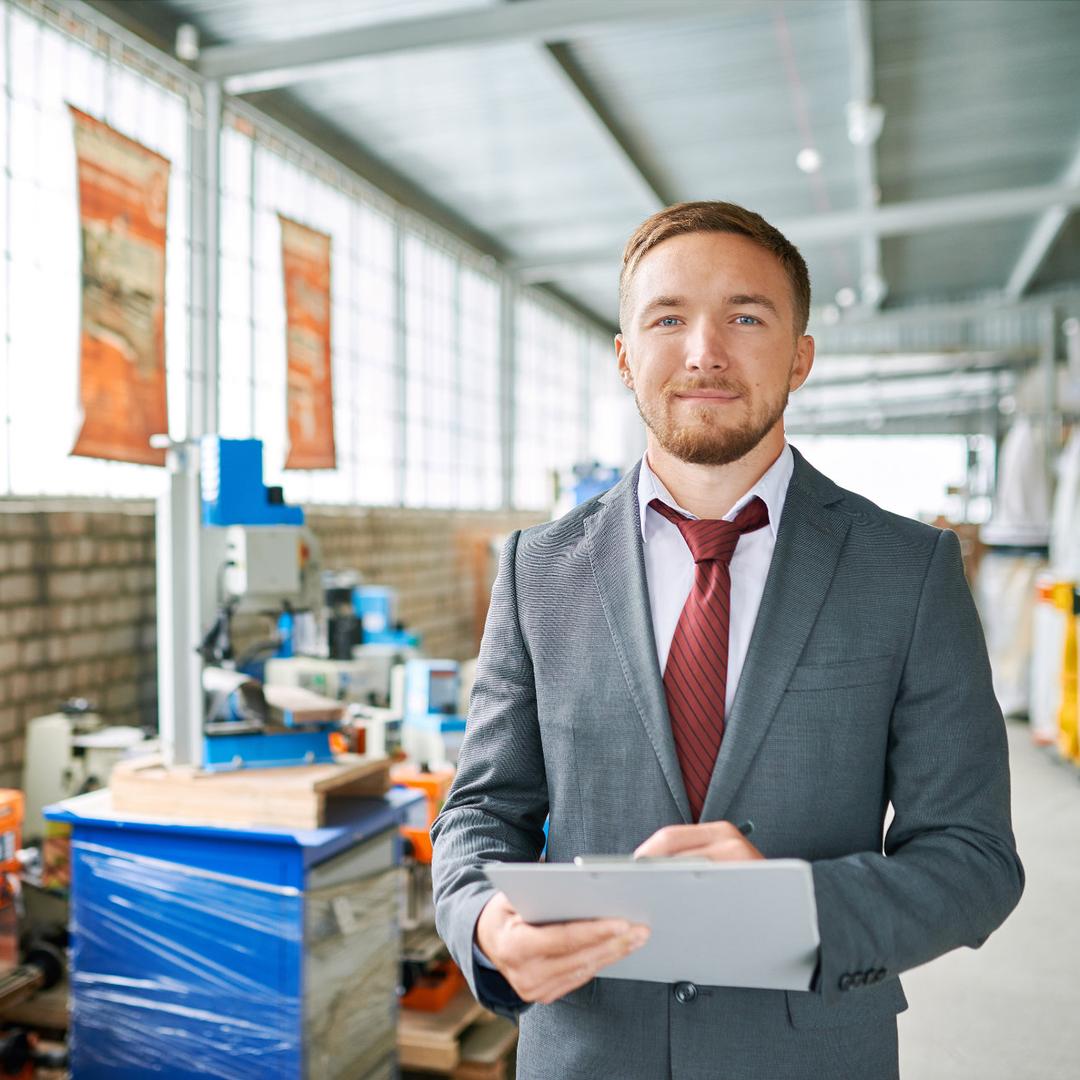 Ein edel gekleideter Mann steht in einer Industriehalle mit einem Klemmbrett in der Hand und schaut lächelnd in die Kamera.