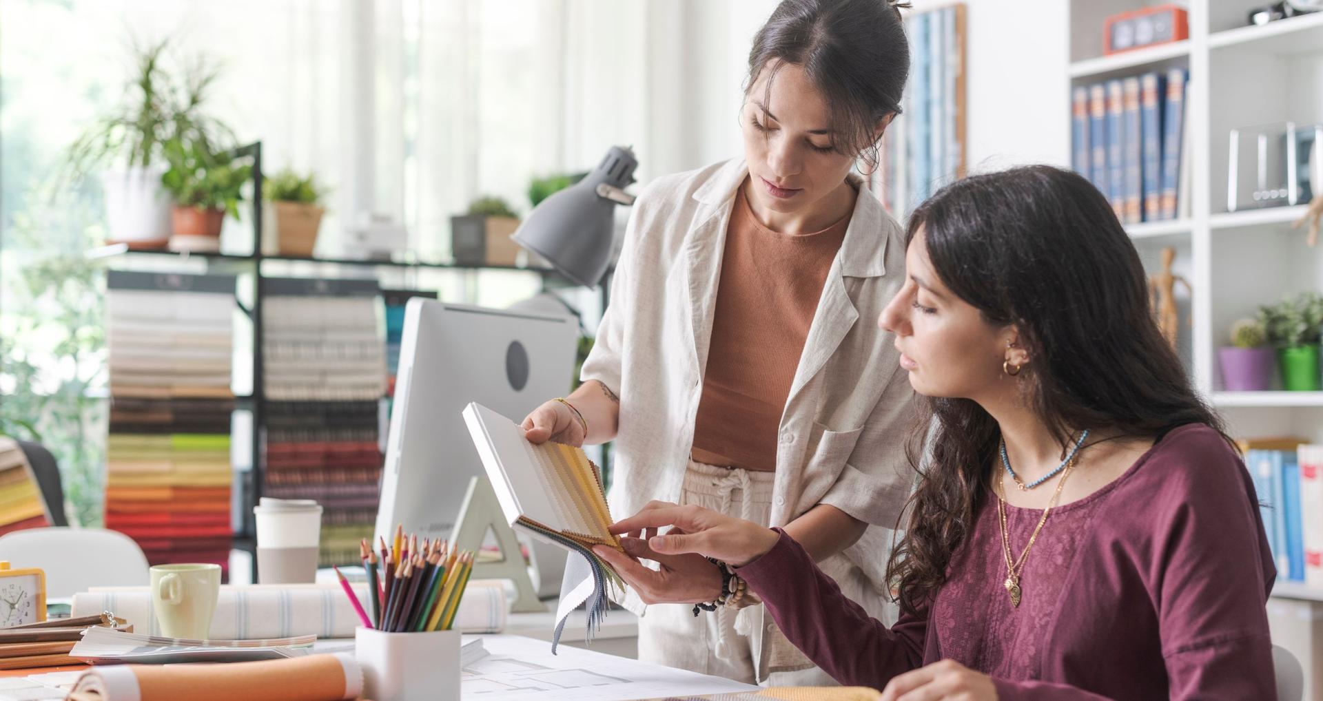 Zwei junge Frauen in einem hellen Innenarchitektur-Studio betrachten gemeinsam ein Musterbuch mit Stoffproben. Im Hintergrund sind Regale mit Farbfächern, Pflanzen und Büchern zu sehen.