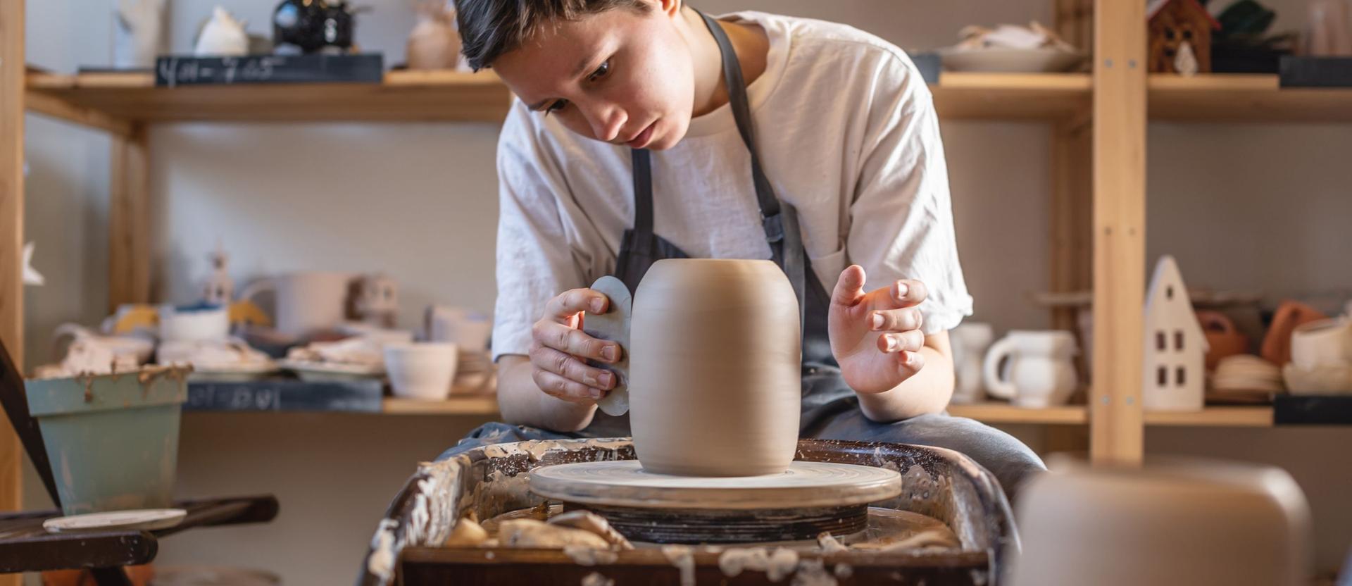 Eine Frau töpfert eine Vase und streicht sie mit einem bestimmten Tool glatt. Sie sitzt dabei auf einem Hocker in einer Töpfer-Werkstatt.