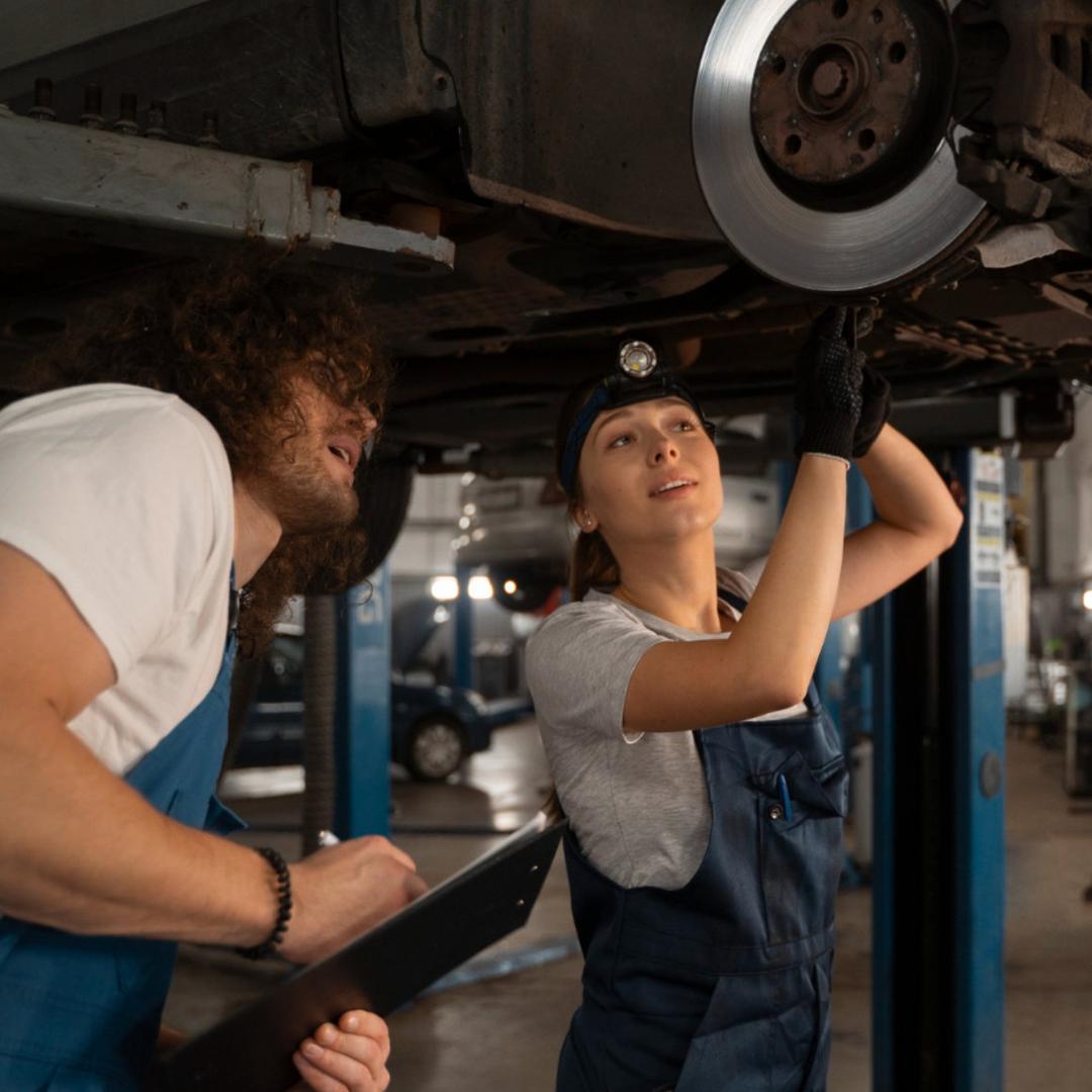 Ein Kfz-Mechatroniker und eine Kfz-Mechatronikerin in Arbeitskleidung, schauen sich gemeinsam etwas unter einem Auto an.