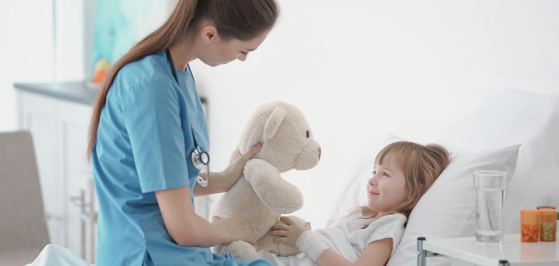 Eine junge Pflegerin besucht ein kleines Mädchen im Krankenzimmer. Sie bringt das Kind mit einem Stofftier zum Lächeln.