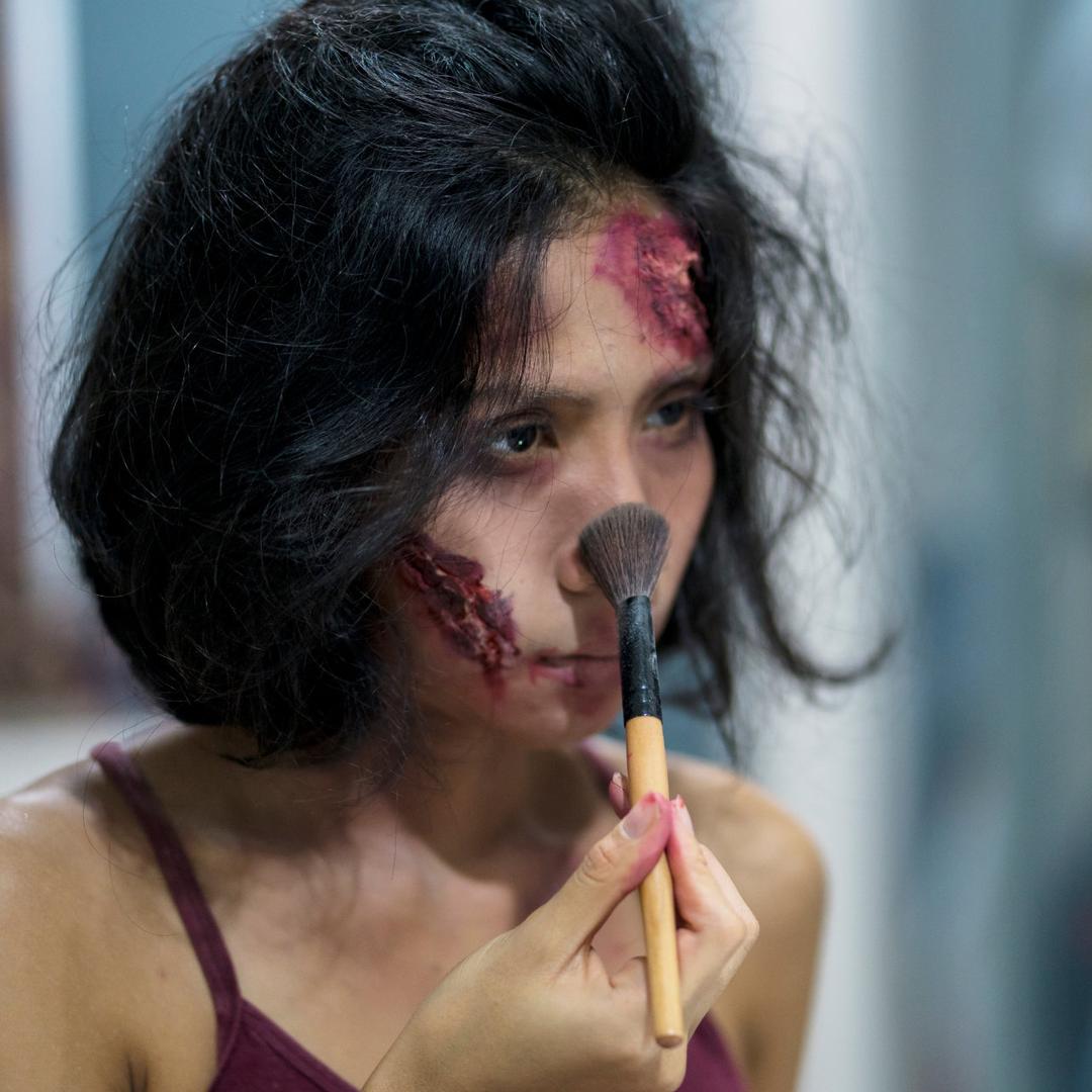 Eine junge Frau schaut ernst in den Spiegel, während sie sich ein aufwendiges Bühnen-Make-Up schminkt.