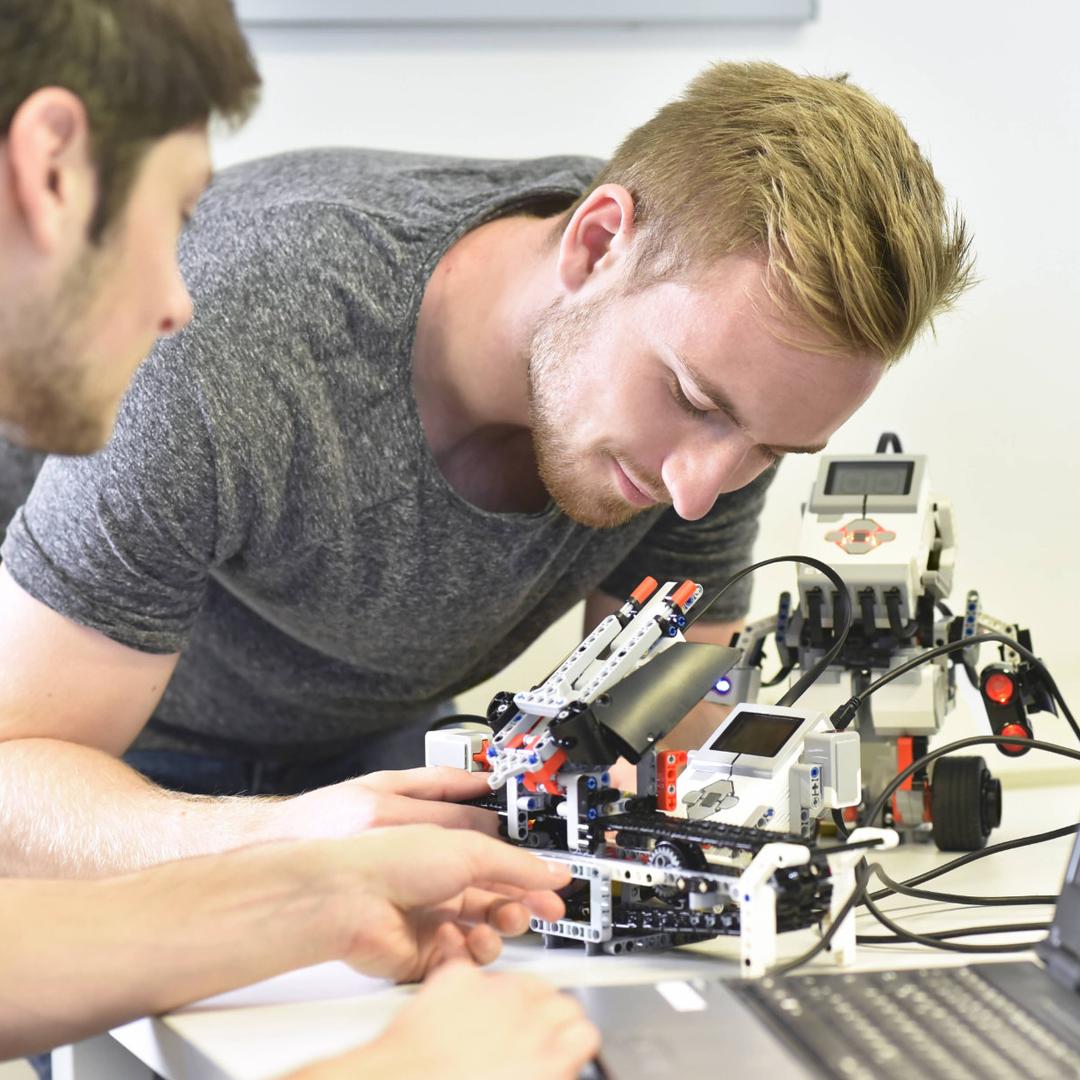 Zwei Personen arbeiten gemeinsam an einem technischen Projekt und überprüfen Bauteile an einem Robotik‑Modell neben einem aufgeklappten Laptop.