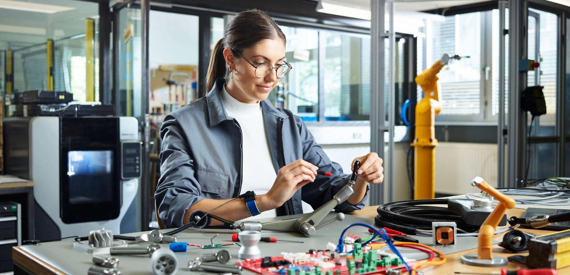 Eine junge Frau, vermutlich eine Mechatronikerin, arbeitet mit technischen Bauteilen in einer Industriewerkstatt.