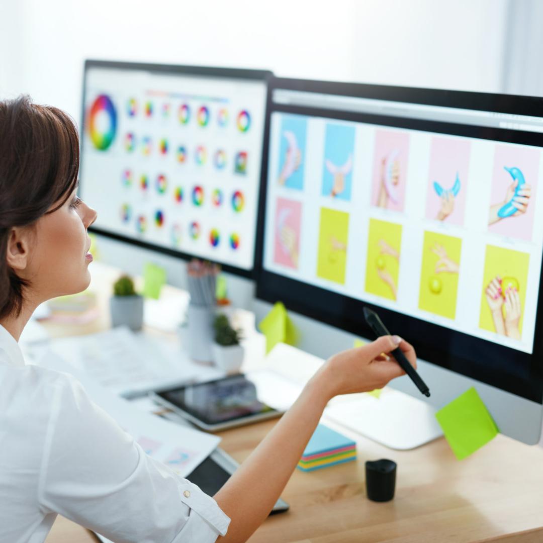 Zwei Personen sitzen an einem Schreibtisch und arbeiten an großen Monitoren, auf denen farbenfrohe Grafikdesign-Entwürfe und Farbräder angezeigt werden.