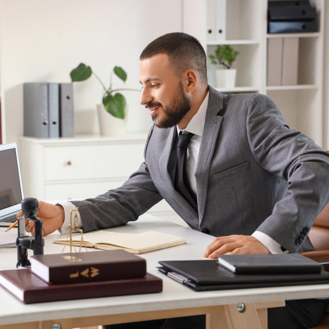 Zwei Männer sitzen in einem Büro am Schreibtisch, während einer im Anzug auf ein Dokument zeigt, das der andere gerade unterschreibt.