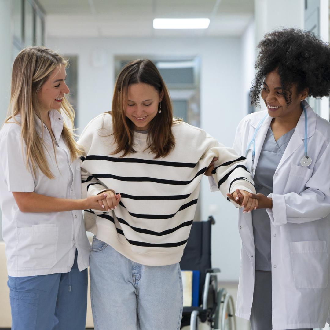 Zwei medizinische Fachkräfte unterstützen eine junge Frau beim Gehen durch einen Krankenhausflur.