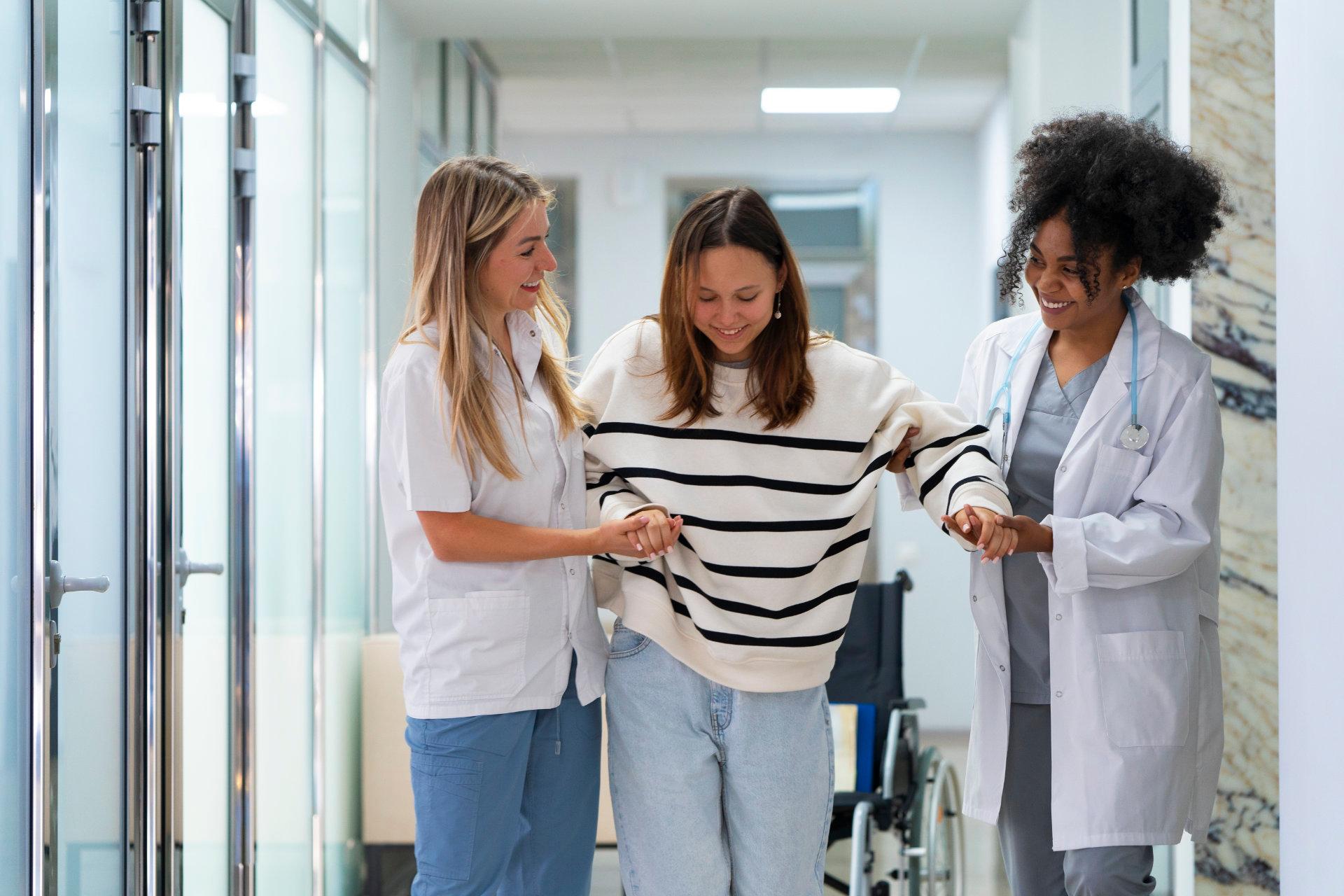 Zwei medizinische Fachkräfte unterstützen eine junge Frau beim Gehen durch einen Krankenhausflur.