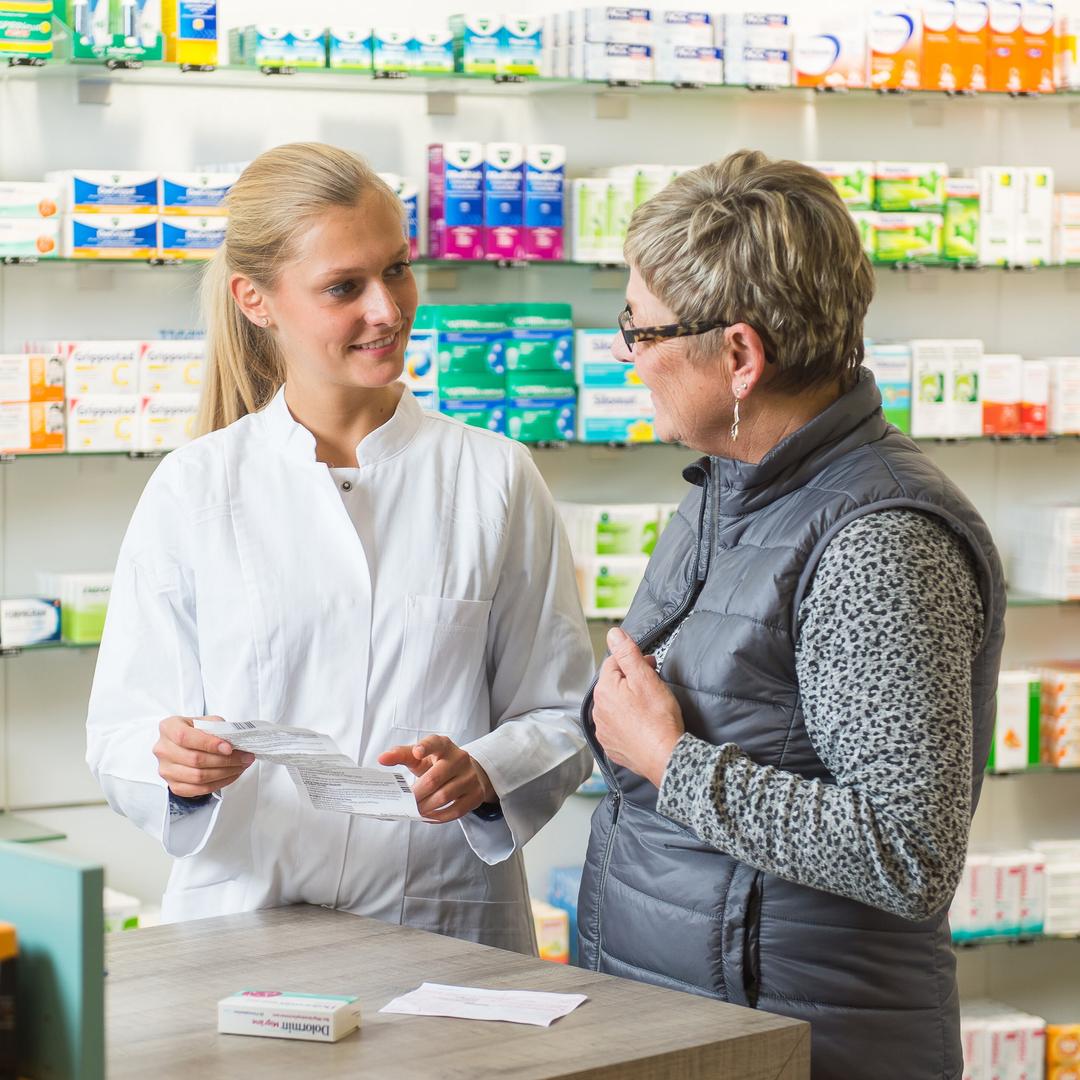 Eine Person im weißen Kittel berät eine Kundin an der Apothekentheke und erklärt ein Medikament, während im Hintergrund gut gefüllte Regale mit Arzneimitteln zu sehen sind.