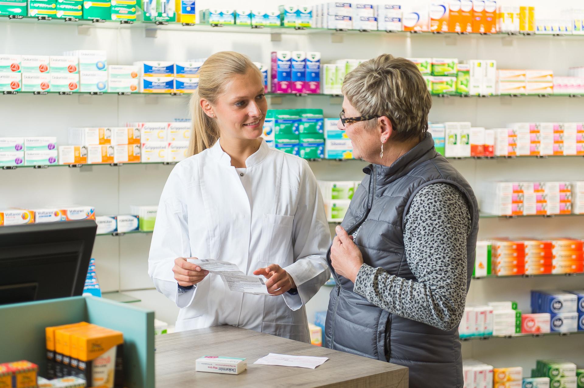 Eine Person im weißen Kittel berät eine Kundin an der Apothekentheke und erklärt ein Medikament, während im Hintergrund gut gefüllte Regale mit Arzneimitteln zu sehen sind.