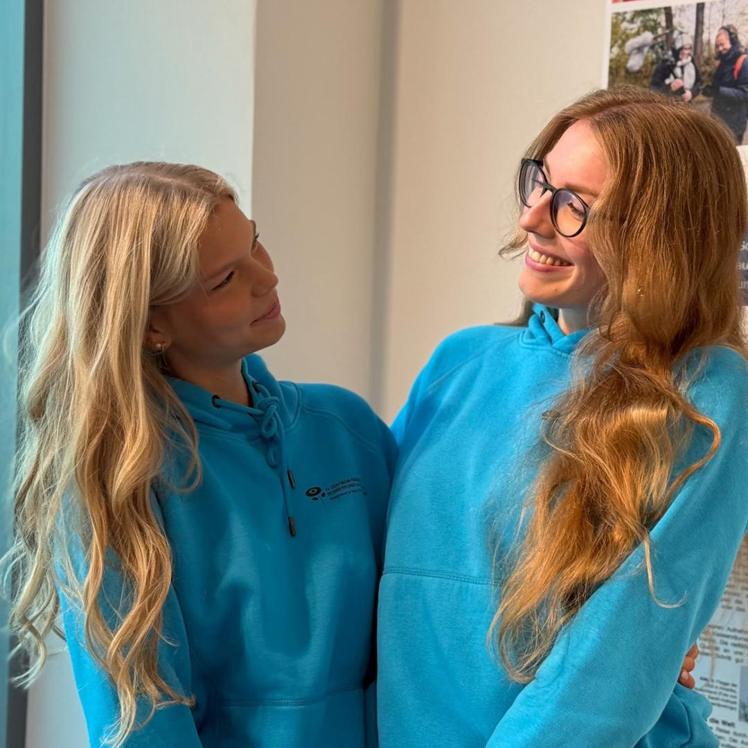 Zwei junge rothaarige Pflegefachfrauen mit Brillen und langen blonden Haaren stehen eng umarmt lachend in einem Flur neben einem Fenster. Sie tragen beide blaue Hoodies und hinter ihnen hängen Poster einer Hochschule.