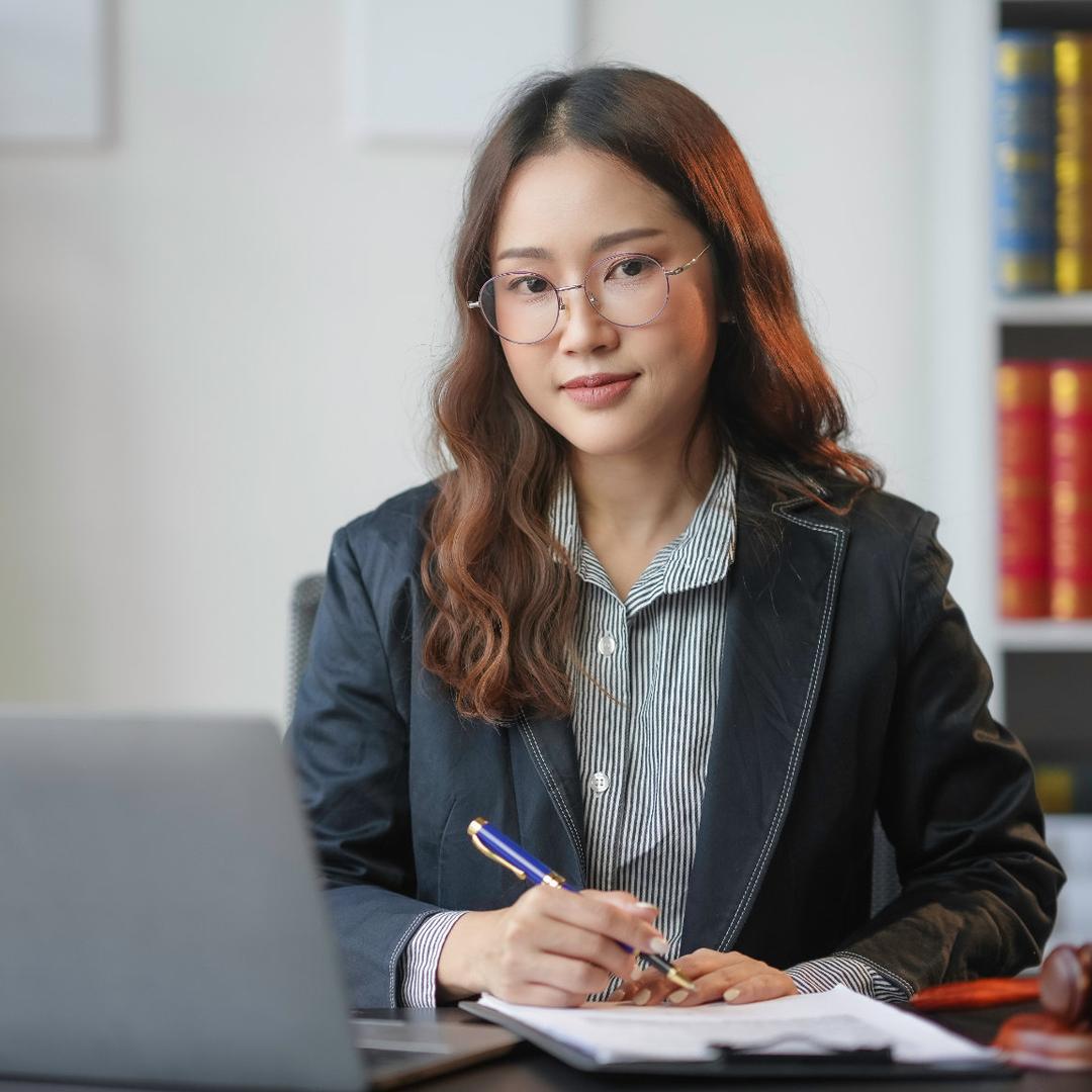 Eine junge asiatische Frau mit Brille und langen welligen Haaren sitzt als Rechtsanwaltsfachangestellte an einem Schreibtisch in einer Kanzlei. Sie trägt eine dunkelblaue Business-Bluse, hält einen Kugelschreiber in der Hand und notiert etwas in ein offenes Notizbuch neben einem Laptop. Im Hintergrund stehen Regale mit Gesetzbüchern, Aktenordnern und zwei goldenen Justitia-Figuren sowie ein weiterer Hammer.