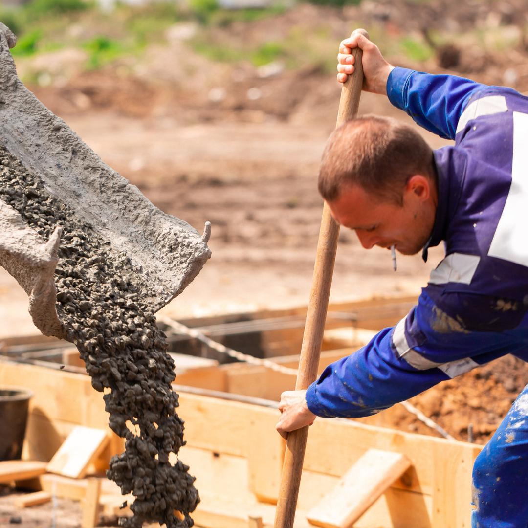 Ein Stahl und Betonbauer in blauer Arbeitskleidung verarbeitet auf einer Baustelle frischen Beton und arbeitet konzentriert mit Werkzeug an einem Fundament.
