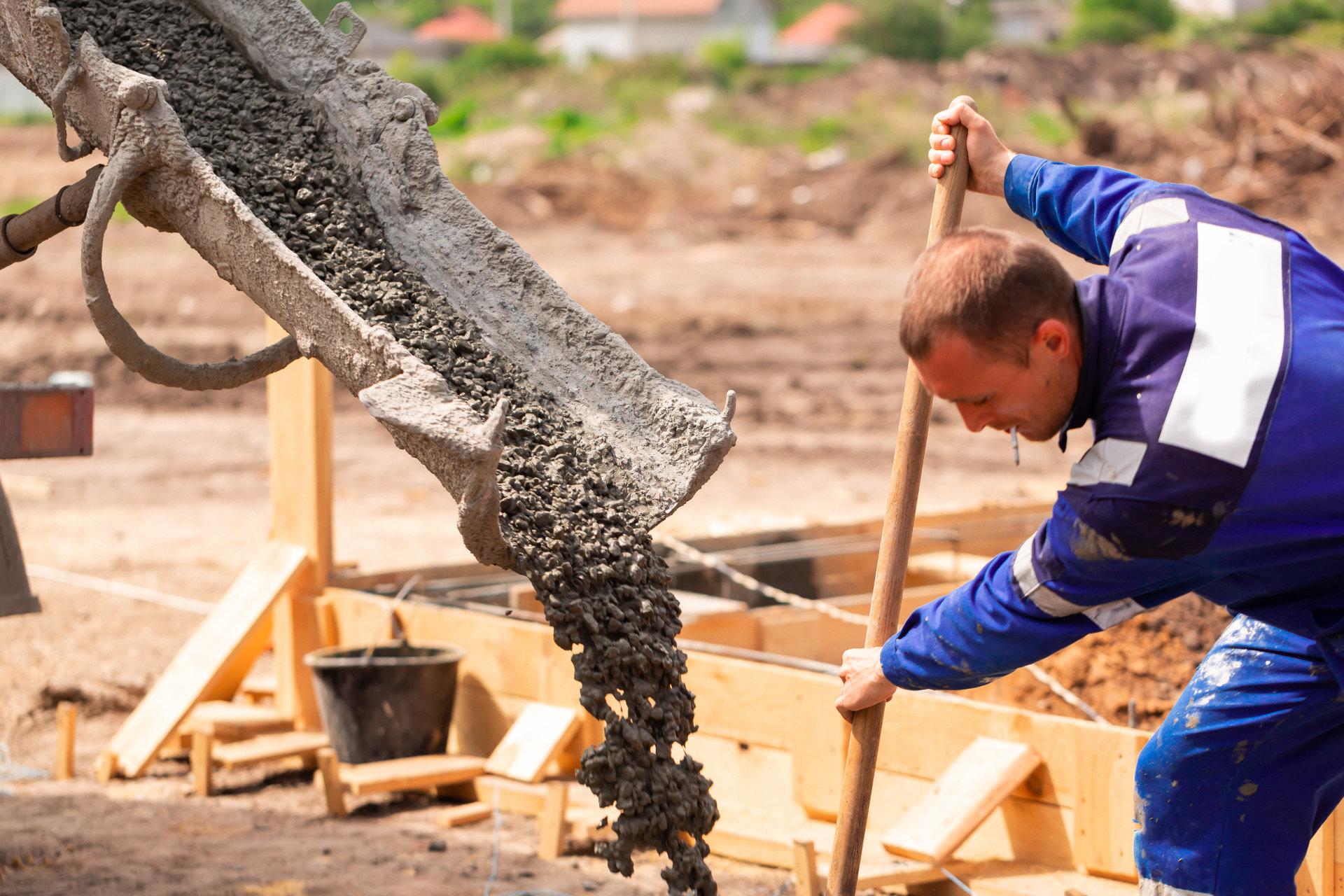 Ein Stahl und Betonbauer in blauer Arbeitskleidung verarbeitet auf einer Baustelle frischen Beton und arbeitet konzentriert mit Werkzeug an einem Fundament.