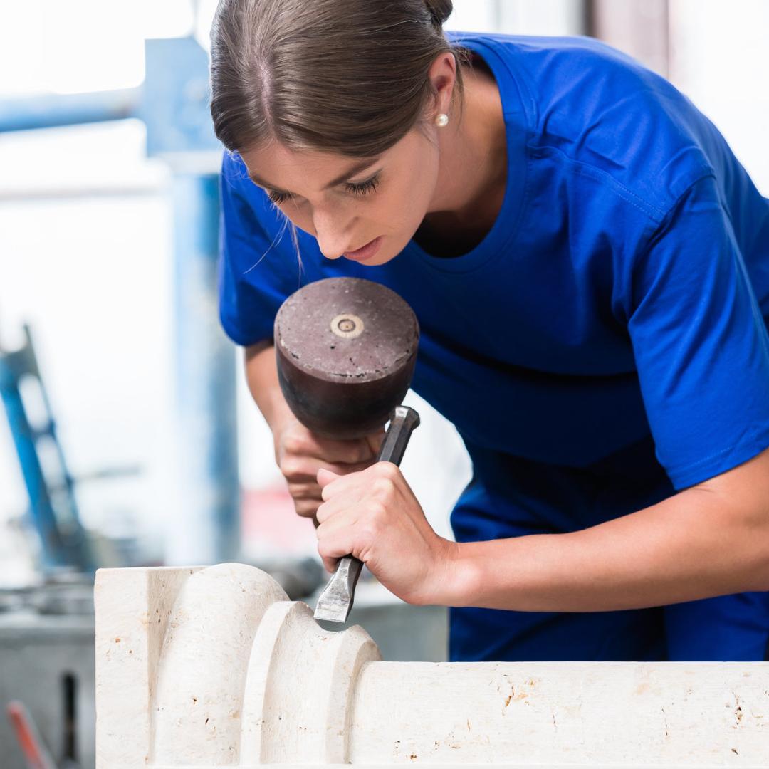Eine Steinmetzin und Steinbildhauerin in blauer Arbeitskleidung bearbeitet mit Hammer und Meißel sorgfältig ein Werkstück aus hellem Naturstein.