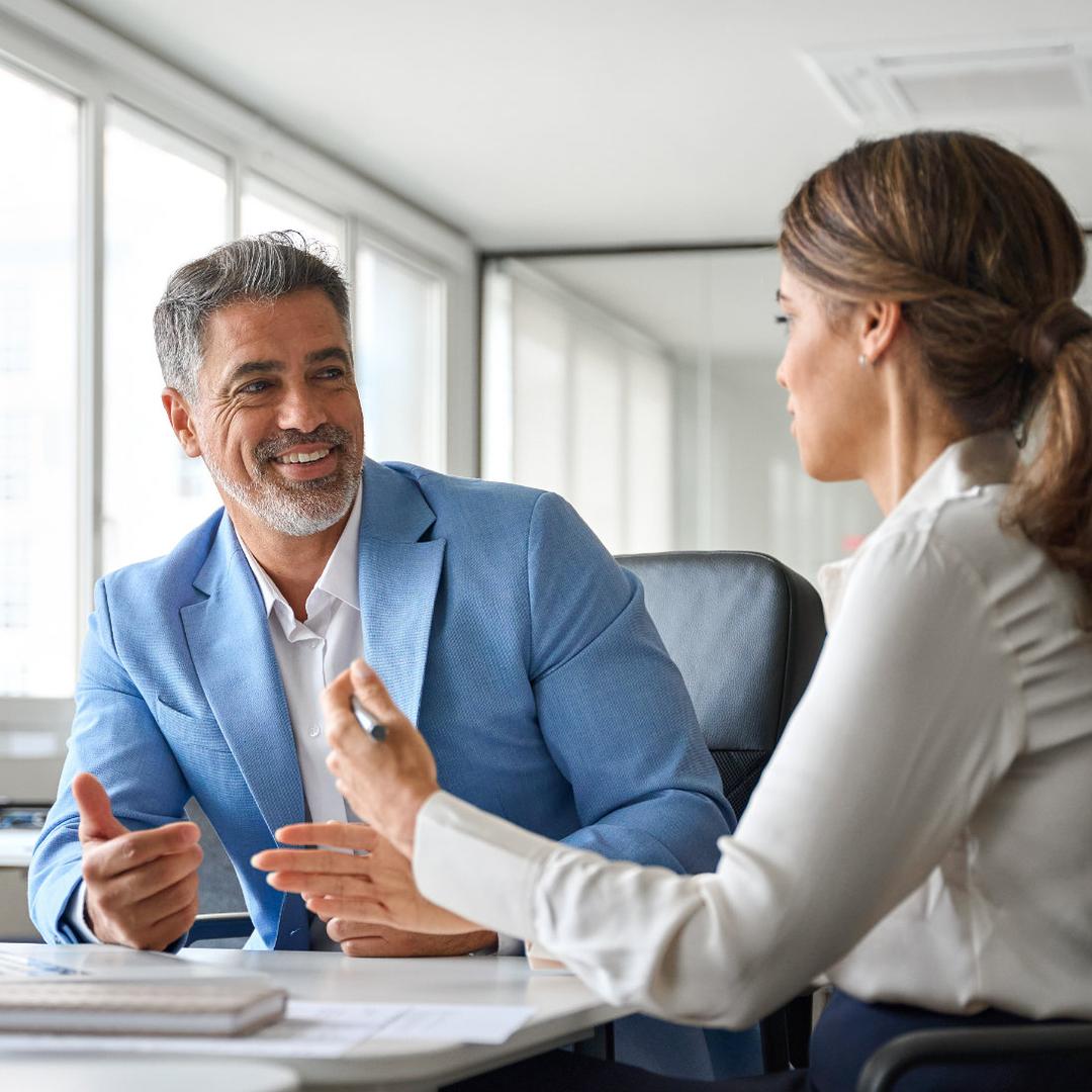 In einem hellen Büro sitzen zwei Personen an einem Tisch und sprechen miteinander; vor ihnen liegen Unterlagen und ein geöffneter Laptop. Eine Person trägt ein hellblaues Sakko, die andere eine weiße Bluse, und beide gestikulieren während des Gesprächs.