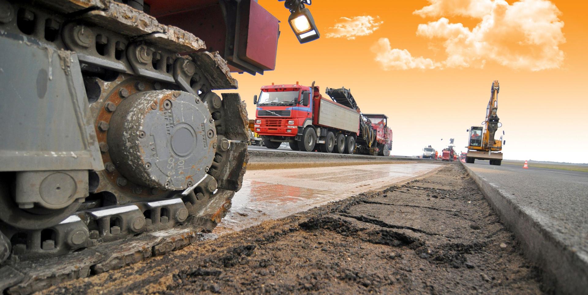 Auf einer Straße fahren verschiedene Baustellenfahrzeuge, wie Bagger, umher und glätten den noch feuchten Beton.