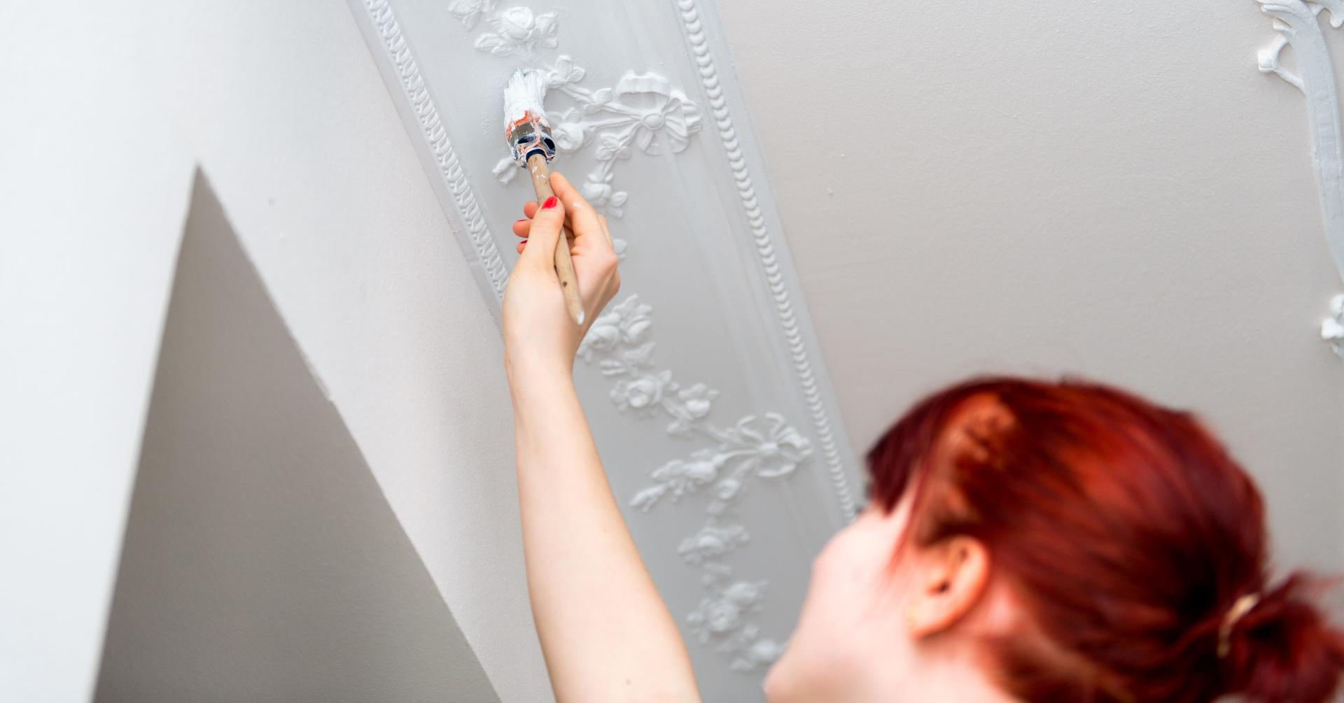 Eine Frau pinselt mit weißer Farbe eine Wand mit Stuck an.