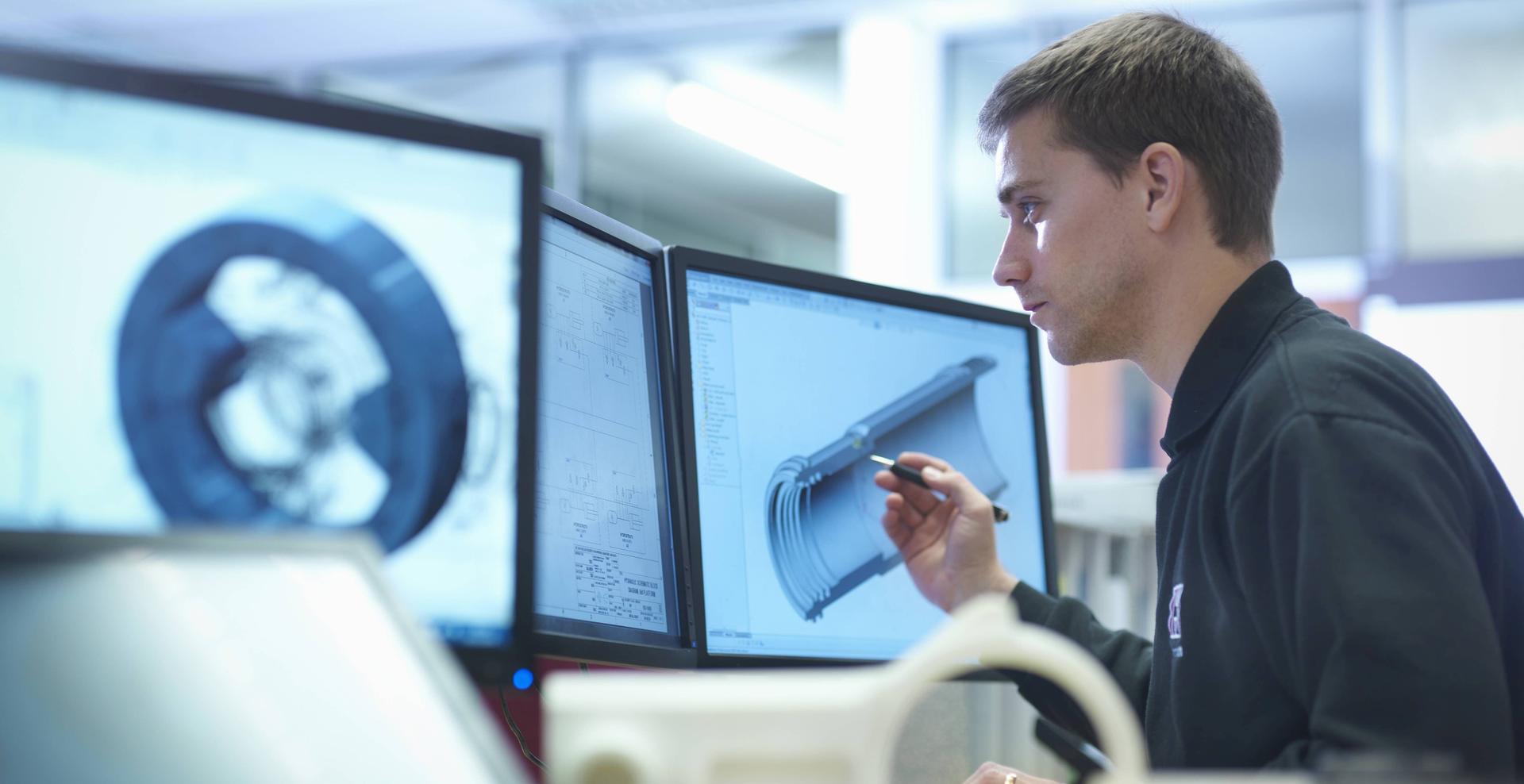 Ein junger Mann, vermutlich ein Technischer Produktdesigner, arbeitet am Computer und erstellt verschiedene digitale 3D-Modelle mit CAD.