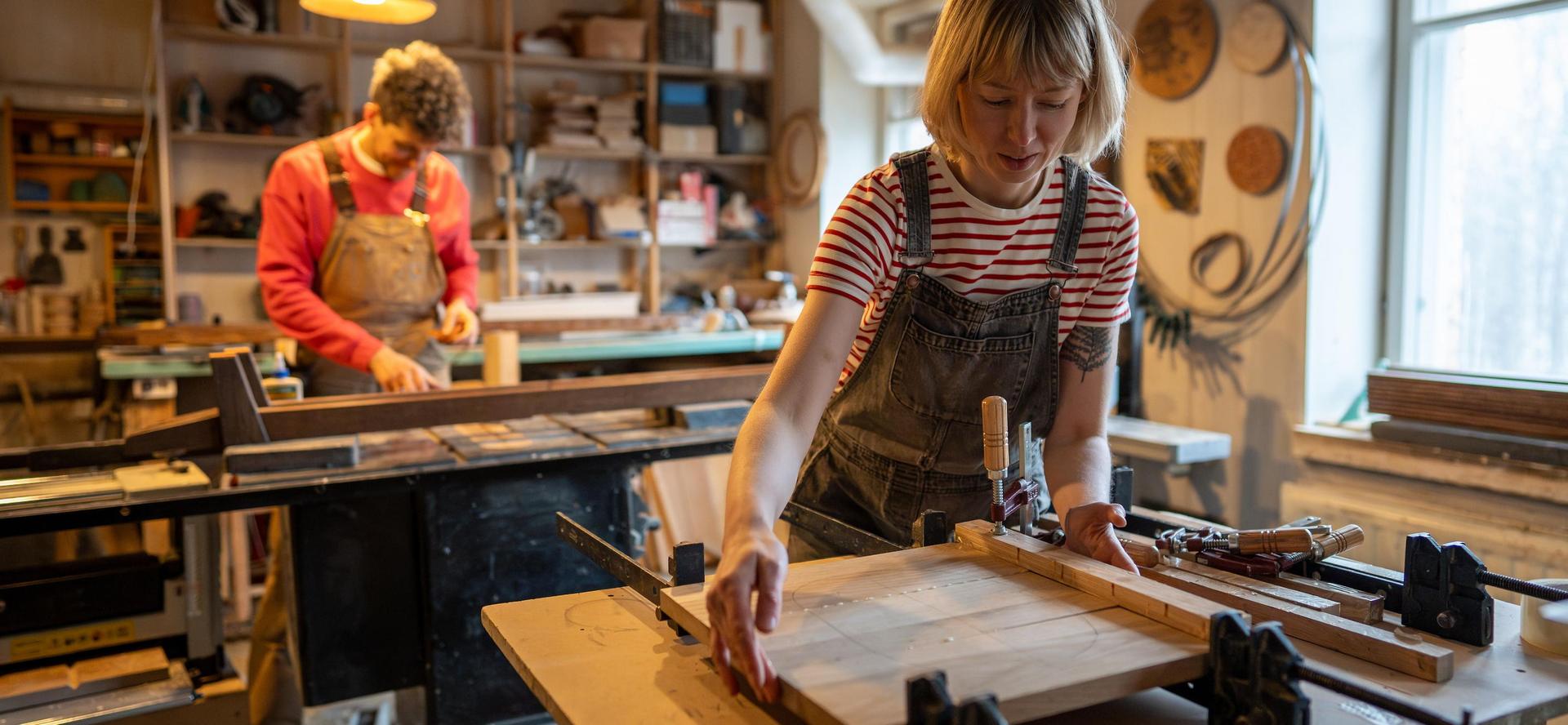 Im Vordergrund steht eine Frau an einer großen Werkbank. Sie trägt eine Latzhose aus Stoff und ein kurzärmeliges, rot-weiß gestreiftes Shirt. Ihre blonden Haare sind kurz geschnitten. Sie beugt sich leicht nach vorne und arbeitet konzentriert an einer Holzplatte. Das Holzstück ist mit Schraubzwingen auf dem Tisch fixiert. Mit der Hand richtet sie das Brett aus oder prüft die Oberfläche. Neben ihr liegen Handwerkzeuge aus Holz und Metall.